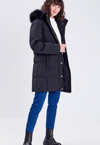 Manteau en doudoune noir avec une capuche en fausse fourrure, comportant des fermetures à boutons, des poches latérales et un design matelassé, porté sur un pantalon bleu à motifs.
