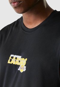 Schwarzes, ärmelloses Shirt mit rundem Ausschnitt, das das weiße und gelbe "Los Angeles Lakers"-Logo auf der Brust trägt. Glatte Stofftextur.