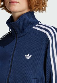 Marineblaue Jacke mit Reißverschluss und strukturierter Strickoptik, mit weißen Drei-Streifen-Akzenten an den Schultern und einem kleinen weißen Logo auf der Brust.