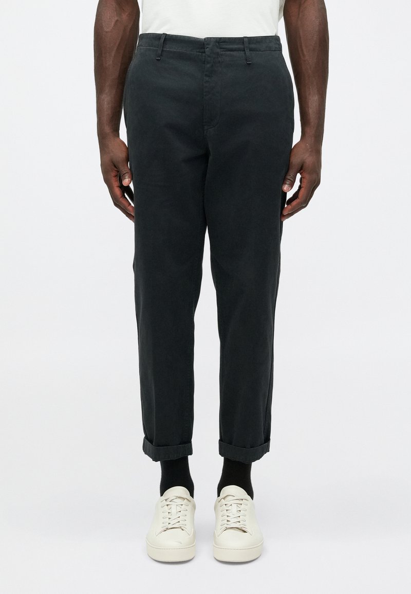 Černé bavlněné chinos kalhoty s tailoring fit, s ohrnutými manžetami, předními kapsami a hladkou texturou, doplněné o bílé tenisky a černé ponožky.
