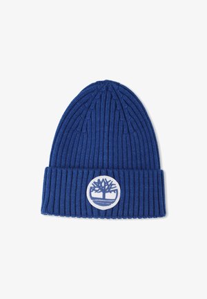 Blauwe geribbelde gebreide beanie met een omslagen manchet en een ronde witte logopatch met boomontwerp. Zachte, gestructureerde stof.