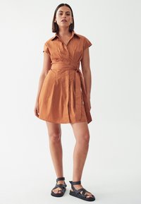 Kurzärmliges, kragenloses Kleid aus rostfarbigem Stoff mit gebundenem Taillenzug und schwingendem Rock. Kombiniert mit schwarzen Riemchensandalen.