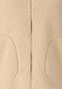 Close-up af beige sherpa-fleece-stof med synlig lynlås og to runde laplommer på et beklædningsstykke.