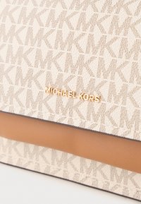 Michael Kors kott, millel on beezist kangast väliskate, millel on pruuni nahast detailid ja kullatud logo ettepoole.