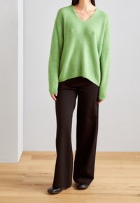 Pull vert en tricot avec col en V, coupe décontractée et épaules tombantes, associé à un pantalon large noir et des chaussures noires.