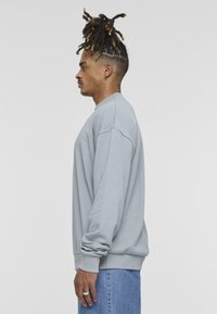 Grå sweatshirt med avslappet passform, ribbestrikket krage og mansjetter, myk tekstur, kombinert med blå denimjeans. Sidesprofilvisning.