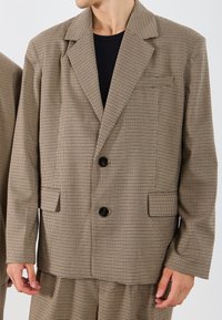 Blazer în carouri maro cu rever căptușit, două buzunare laterale și două nasturi negri. Purtați peste o cămașă neagră. Are o croială lejeră.