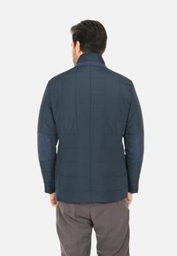 Giacca trapuntata blu navy con colletto alto, dotata di toppe azzurre a contrasto sui gomiti e una superficie liscia.