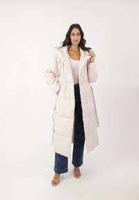 Abrigo acolchado de color rosa claro con capucha, botones a presión y un diseño largo. Combinado con jeans de mezclilla oscuros y sandalias de tacón transparente.