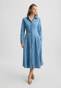 Hellblaues Denim-Hemdkleid mit langen Ärmeln, Knopfverschluss vorne, tailliertem Schnitt und weit schwingendem Rock. Musterhafte Schuhe vervollständigen das Outfit.