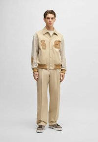 Beżowa kurtka varsity z beżowymi rękawami i haftowanymi naszywkami. Połączona z beżowymi spodniami o szerokich nogawkach i szarymi trampkami. Gładka tekstura.