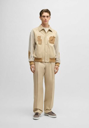 Beżowa kurtka varsity z beżowymi rękawami i haftowanymi naszywkami. Połączona z beżowymi spodniami o szerokich nogawkach i szarymi trampkami. Gładka tekstura.