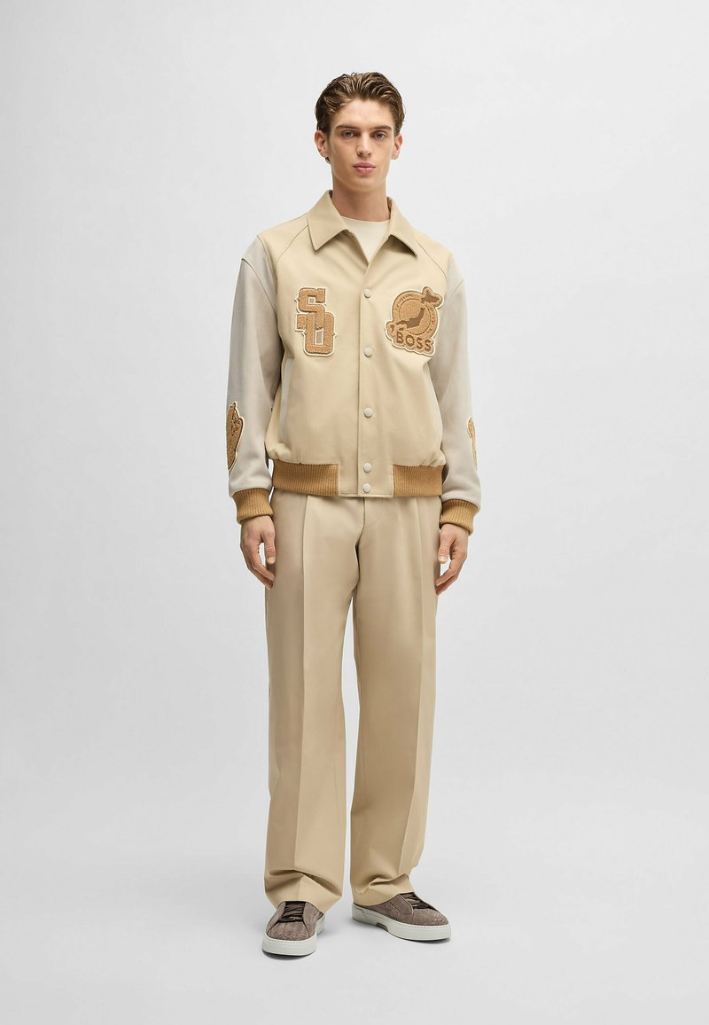 Beżowa kurtka varsity z beżowymi rękawami i haftowanymi naszywkami. Połączona z beżowymi spodniami o szerokich nogawkach i szarymi trampkami. Gładka tekstura.
