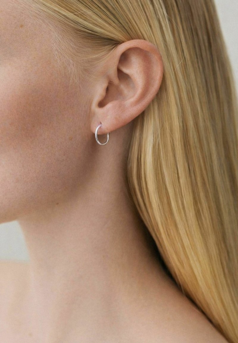 Gros plan sur l'oreille gauche d'une personne portant une petite boucle d'oreille en argent, encadrée par des cheveux blonds et raides sur un fond neutre.
