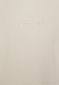 Logo Adidas na světle béžové tkanině, s jemně vyšitým designem. Hladká textura s minimalistickou estetikou.