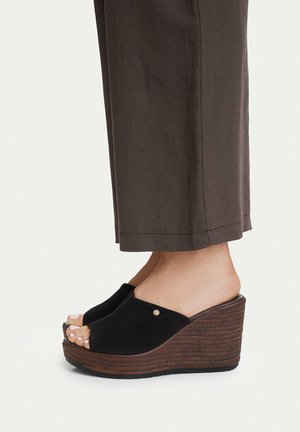 Piedi con sandali con zeppa in camoscio nero e punta aperta con plateau in legno, abbinati a pantaloni marroni corti su sfondo semplice.