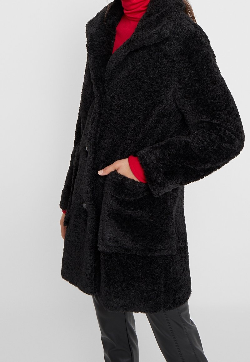 Svart fuskpälsjacka med raggkrage, knäppning framtill och två stora fickor. Texturen är mjuk och fluffig.