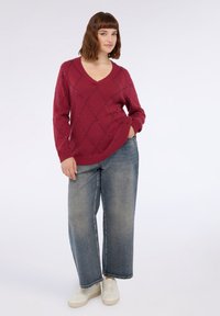 Pulover tricotat maro cu guler în V, având un model în diamant cu broderii. Asortat cu blugi largi de un albastru deschis și adidași albi.