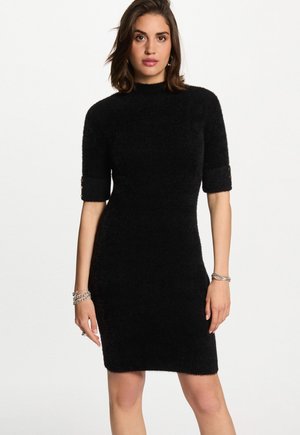 Donna che indossa un vestito a maglia nera aderente con maniche di lunghezza media e braccialetti d'argento a entrambi i polsi, in piedi su uno sfondo bianco semplice.