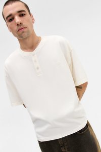 Off-white short-sleeve henley shirt with a round neckline and five-button placket. Features a subtle logo on the chest. Soft material.