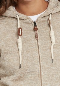 Sudadera de punto beige con cremallera, cuerdas gruesas color crema, ojales de metal y tirador de cremallera de cuero, llevada sobre una camisa blanca.