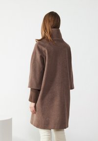 Manteau marron surdimensionné avec un col haut, présentant une texture lisse et des manches larges, porté sur un pull brun côtelé et un pantalon crème.