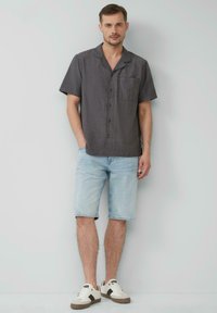 Kurzärmliges, dunkel gemustertes Hemd mit Kragen und Brusttasche, kombiniert mit hellblauen Denim-Shorts und beigen Sneakers.