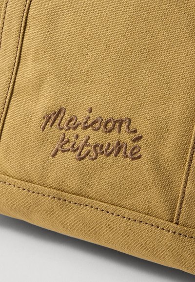 Geltona drobė su rudai siuvinėtu "Maison Kitsuné" logotipu. Turi tekstūruotą paviršių ir matomas siūles palei kraštus.