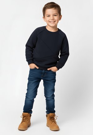 Uśmiechnięty młody chłopiec z rękami w kieszeniach, ubrany w czarną bluzę, niebieskie dżinsy i beżowe sznurowane buty, stoi na jednolitym jasnym tle.