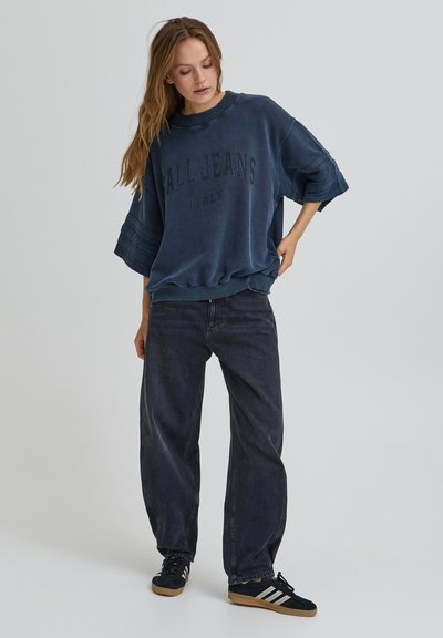 Jeune femme portant un jean ample foncé, un sweat-shirt oversize bleu marine avec le texte "ALL JEANS ITALY", et des baskets noires à rayures blanches.
