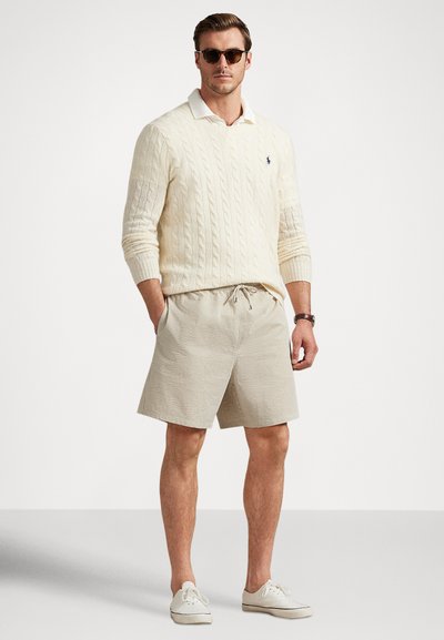 Camisola creme com padrão de malha grossa, camisa branca com gola, calções xadrez bege e ténis claros, complementados com óculos de sol escuros.