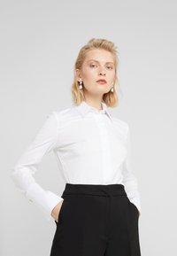 Chemise blanche à manches longues avec une coupe ajustée, col pointu et patte de boutonnage, accompagnée de pantalons noirs taille haute. Texture lisse.