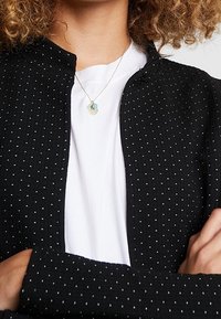 Czarna kurtka o teksturowanej powierzchni w białe kropki, okrągły dekolt, otwarty przód, noszona na białej koszuli, ozdobiona delikatnym naszyjnikiem z kolorowymi charmsami.