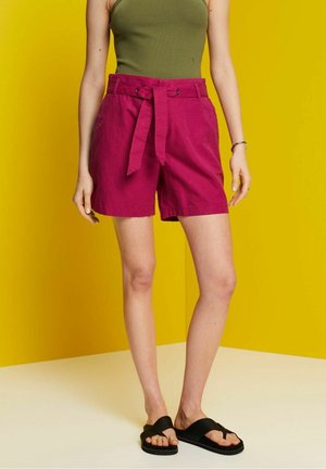 Pink linen shorts with a belted waist, side pockets, and casual fit, paired with black flat sandals. Background is a bright yellow wall.