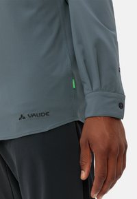 Maglietta grigia a maniche lunghe con texture strutturata, dotata di un logo nero e polsini con bottone. Include un'etichetta verde sulla cucitura laterale.