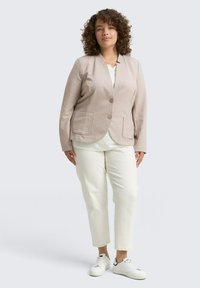 Blazer beige claro con dos bolsillos frontales, cierre de un solo botón, combinado con una blusa blanca y pantalones crema ajustados. Zapatillas blancas completan el look.
