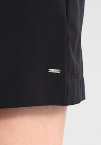 Svarta shorts med en slät textur, prydda med en liten logotagg med röda och blå accenter nära nederkanten. Enkel, funktionell design.