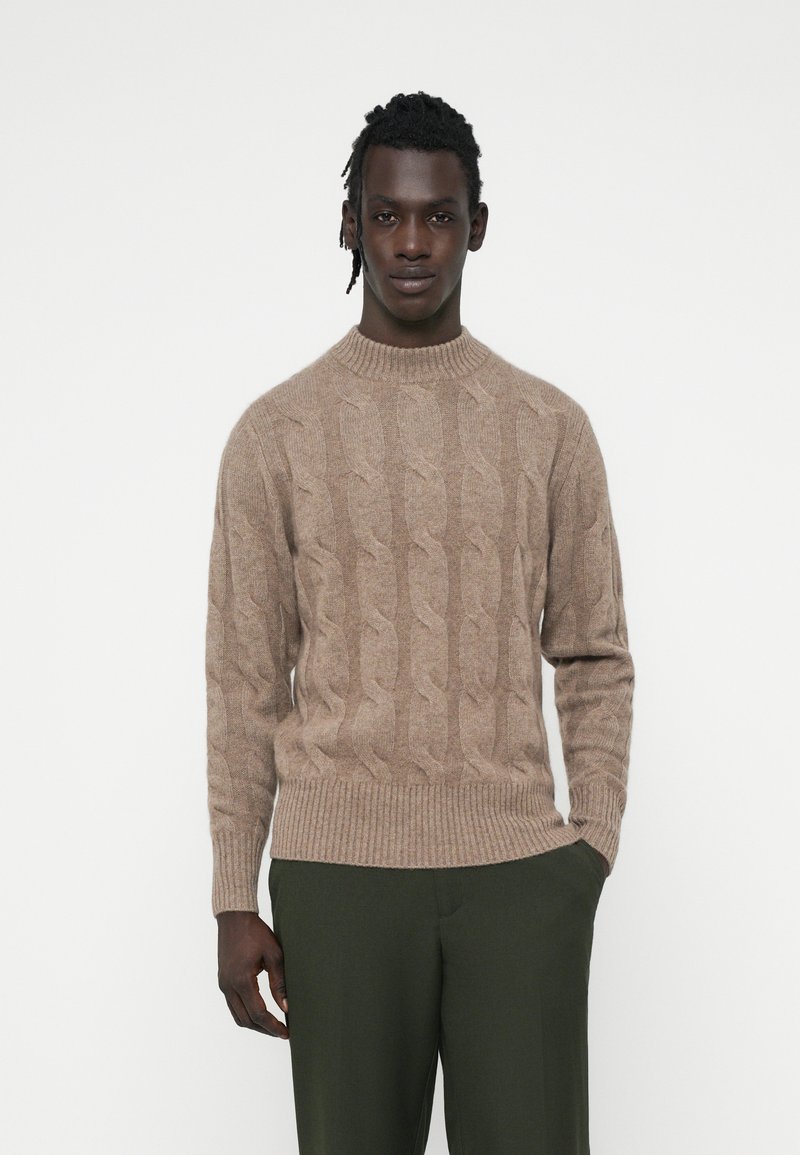 Beige gestrickter Pullover mit hohem Kragen und gerippten Bündchen, kombiniert mit dunkelgrünen Hosen. Weicher Stoff, klassisches Design.