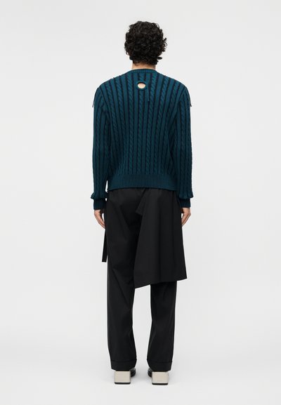 Personne aux cheveux bouclés foncés portant un pull en maille torsadée bleu sarcelle avec une petite ouverture circulaire dans le haut du dos, un pantalon large noir et des chaussures blanches.