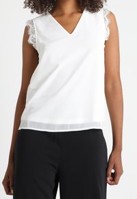 Débardeur blanc sans manches avec un décolleté en V et des accents en dentelle sur les coutures des épaules, présentant un tissu lisse et une coupe décontractée.