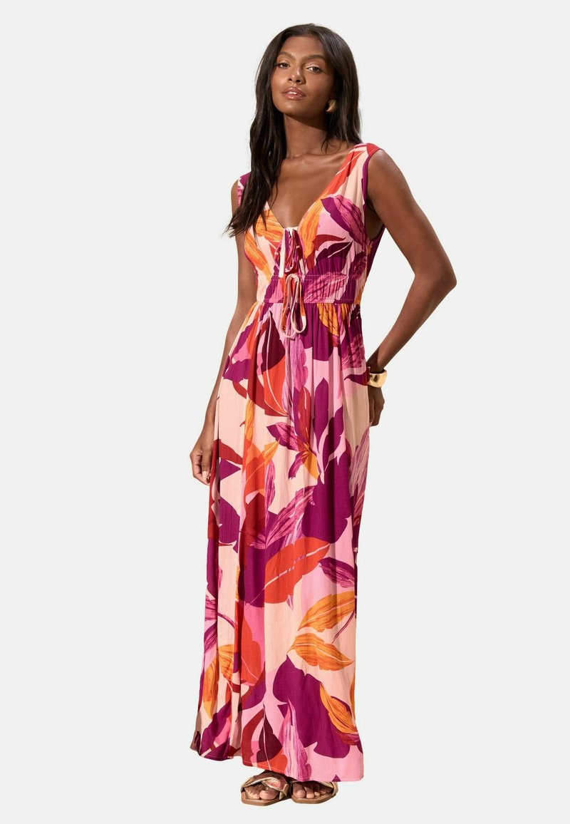 Kvinde iført ærmeløs, lang blomsterkjole i pink, lilla og orange toner, stående med den ene hånd på hoften, iført gyldne sandaler og armbånd.