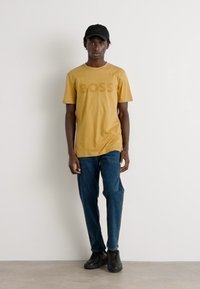 Mustard yellow short-sleeve T-shirt with "BOSS" printed in a lighter shade, paired with blue jeans and black sneakers, worn with a black cap.