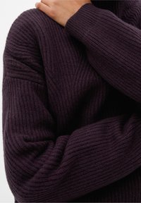 Personne portant un pull côtelé violet foncé qui s'enlace avec une main sur l'épaule opposée, gros plan sur le haut du corps.