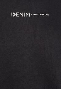 Čierna tkanina s jemnou textúrou, na ktorej je výrazne zobrazený biely potlačený text "IDENIM TOM TAILOR". Jednoduchý, minimalistický dizajn.