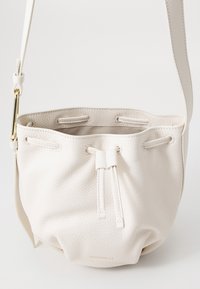 Bílá kůže kbelíková taška s texturovaným povrchem, nastavitelným popruhem, centrálním stahovacím uzávěrem a nenápadným zlatým logem na spodní straně.