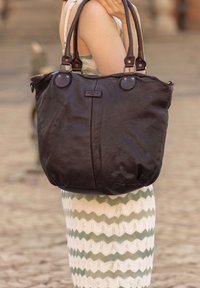 Tamsiai rudos odos rankinė su dviem petnešomis, turinti minkštą tekstūrą ir apvalią formą, ant priekinės pusės yra subtilus logotipas.