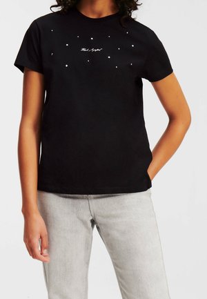 T-shirt noir à manches courtes avec de petits pois blancs et un texte en écriture cursive, associé à un jean gris clair, porté par une personne une main dans la poche.