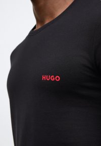 Černé tričko z bavlny s kulatým výstřihem, s červeným logem „HUGO“ na levé straně hrudníku. Hladká textura.