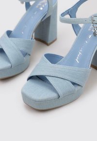 Sandalias de tacón alto de denim azul claro con tiras entrelazadas y un tacón grueso. Textura suave con una parte inferior lisa y ligero plataforma.