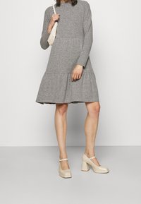 Abito grigio a maniche lunghe con orlo a strati, realizzato in tessuto morbido. Il modello indossa scarpe con tacco a blocco di un leggero beige e tiene una borsa piccola.